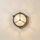 Trident Round Bulkhead Light in antique brass finish, with a three-arm guard over ribbed glass lit from within, mounted on a plain light wall.