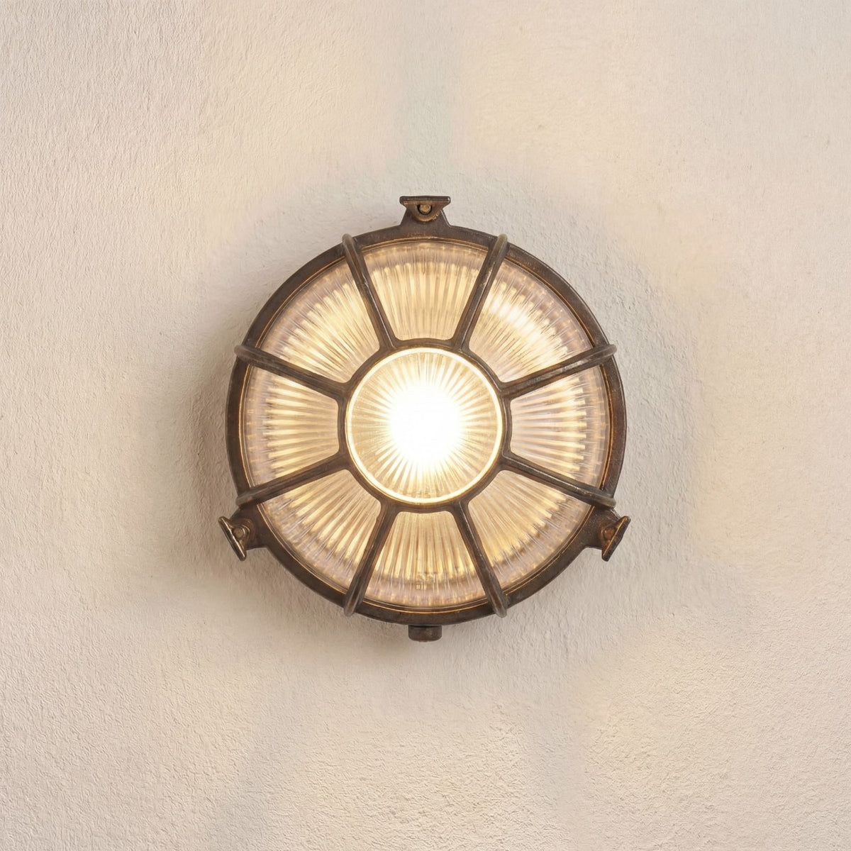 Grand Capstan round bulkhead light in antique brass, with radiating bars over ribbed glass lit from within, mounted on a plain light wall.