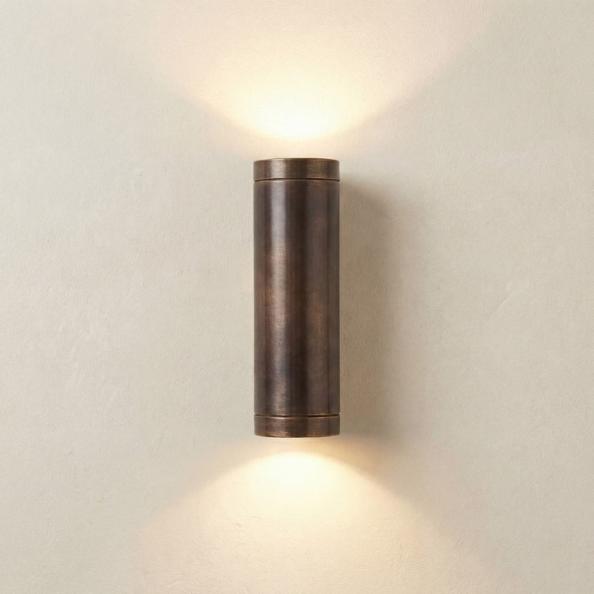 Pharos Outdoor Up & Down Wall Light in antique brass, a slim cylindrical fixture with banded end caps casting light upward and downward on a plain neutral wall.
