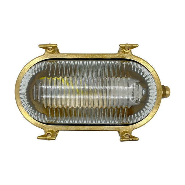 Small Siren oval bulkhead light with solid raw brass body and ribbed clear glass cover, no front grille, shown against a plain white background.