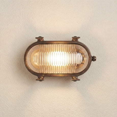 Small Siren oval bulkhead light in antique brass with ribbed glass cover and visible mounting screws, lit on a plain wall background.