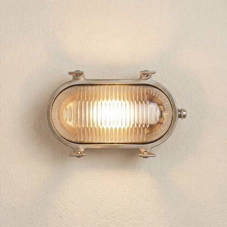 Small Siren Oval Bulkhead Light in satin nickel, solid brass frame with four corner lugs and ribbed glass cover, illuminated on a plain wall background.