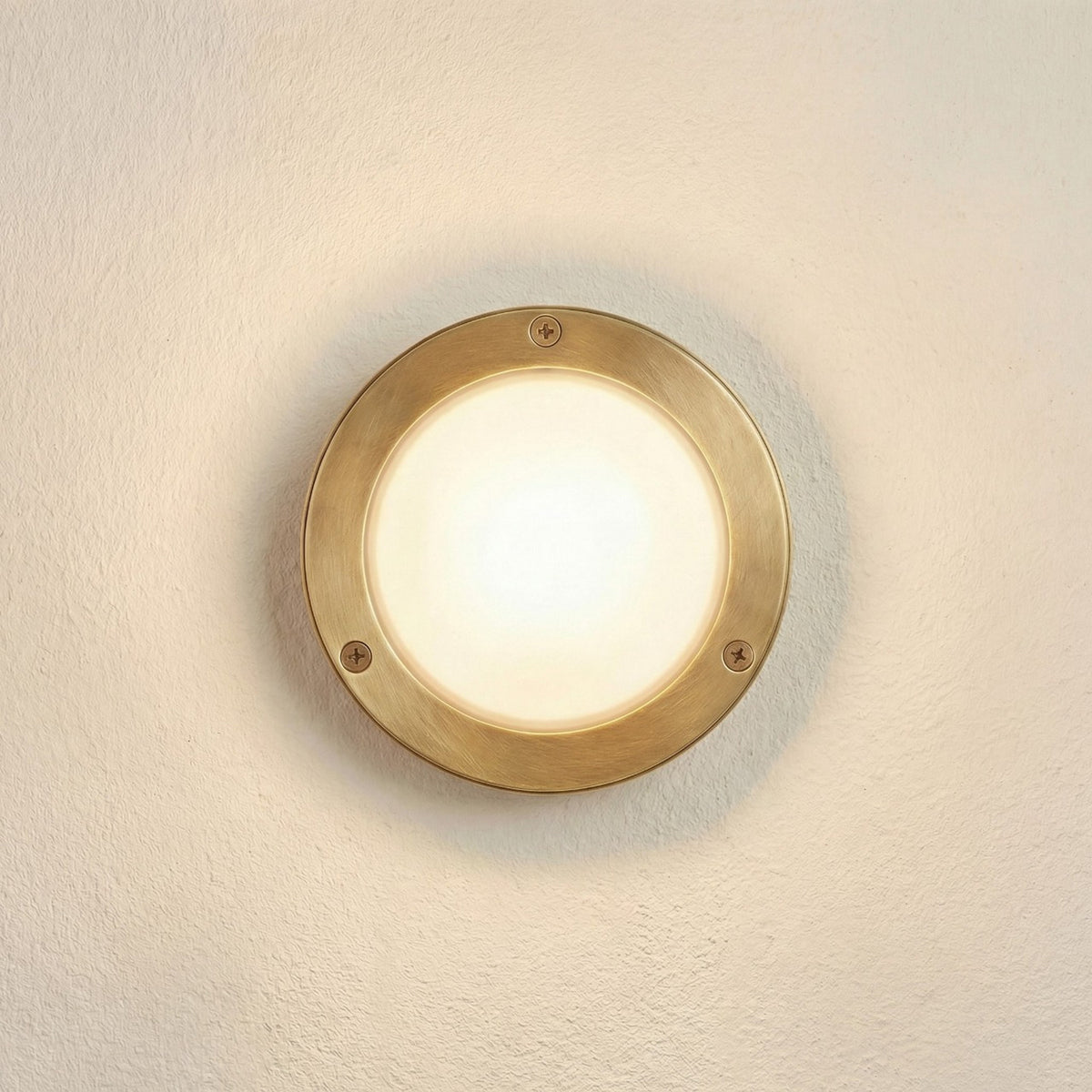 Marina Circular Bulkhead Light IP64 in raw brass, a round wall-mounted fixture with frosted glass dome and three-screw front ring, shown lit on a plain wall.