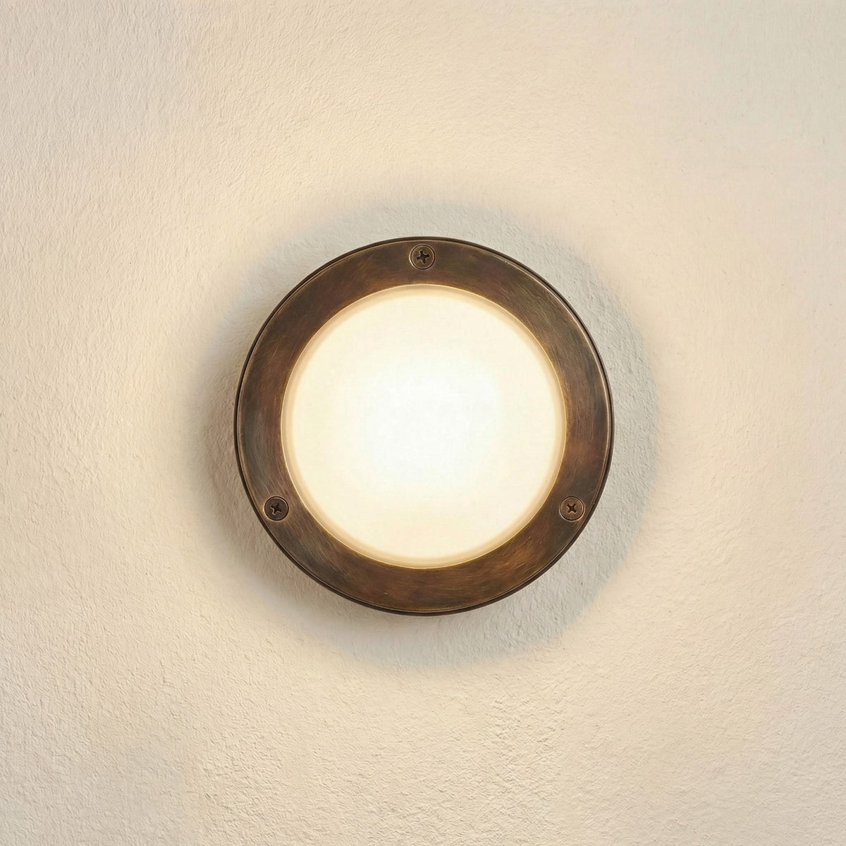 Marina Circular Bulkhead Light in antique brass finish, a round outdoor wall light with a three-screw bezel and domed frosted glass cover, shown illuminated on a plain wall.