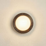 Marina Circular Bulkhead Light in antique brass finish, a round outdoor wall light with a three-screw bezel and domed frosted glass cover, shown illuminated on a plain wall.