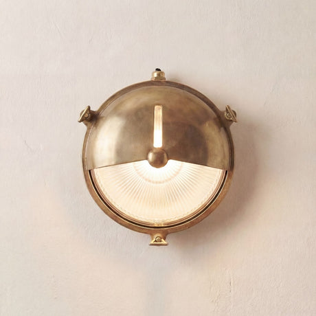 Trident Shield round bulkhead light in raw brass, with a half-moon shield over ribbed glass, slotted top light, and triangular rim lugs, mounted on a plain wall.