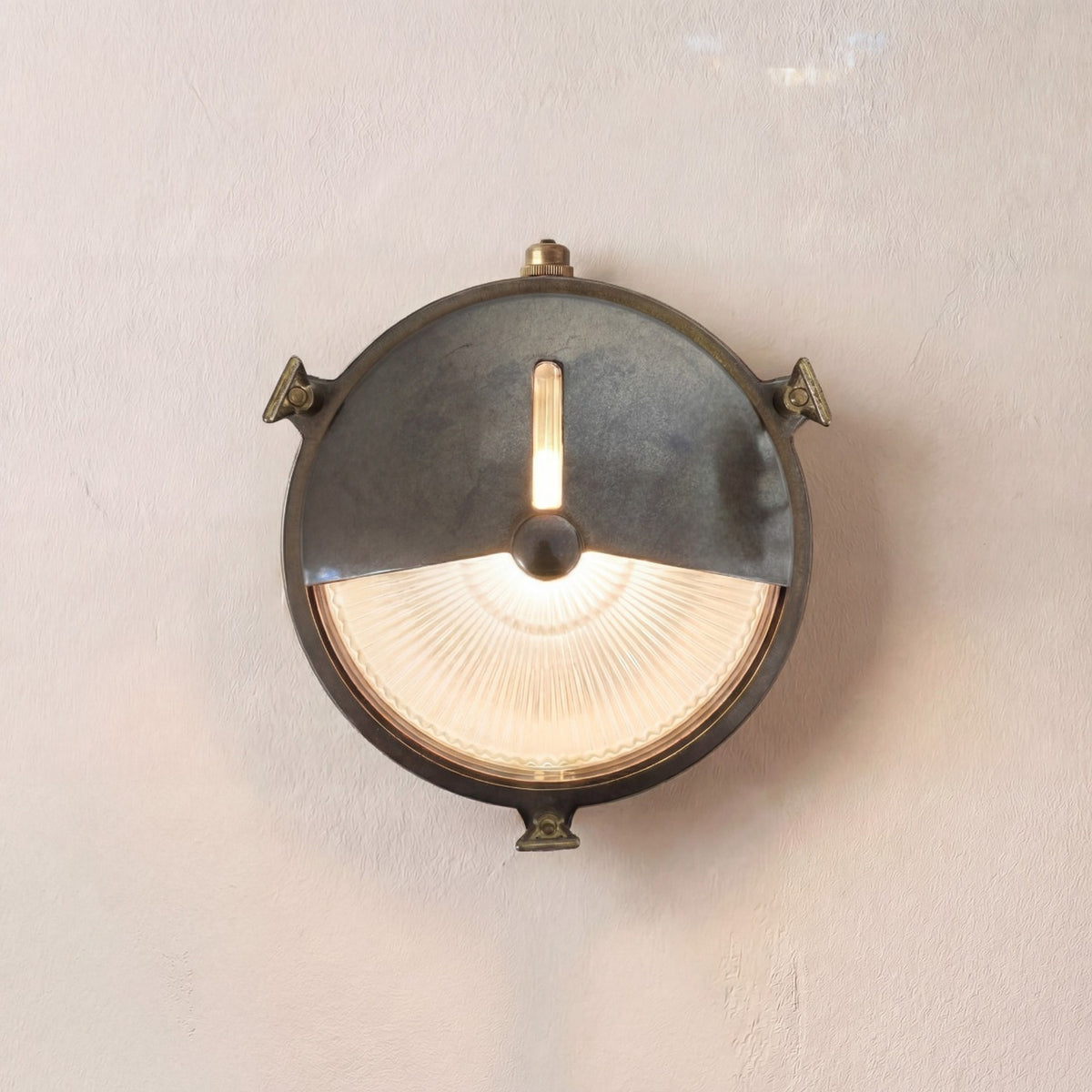 Trident Shield round bulkhead wall light in antique brass with a half shield over ribbed glass, lit bulb visible, mounted on a plain wall.