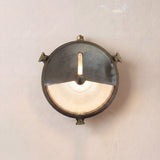 Trident Shield round bulkhead wall light in antique brass with a half shield over ribbed glass, lit bulb visible, mounted on a plain wall.