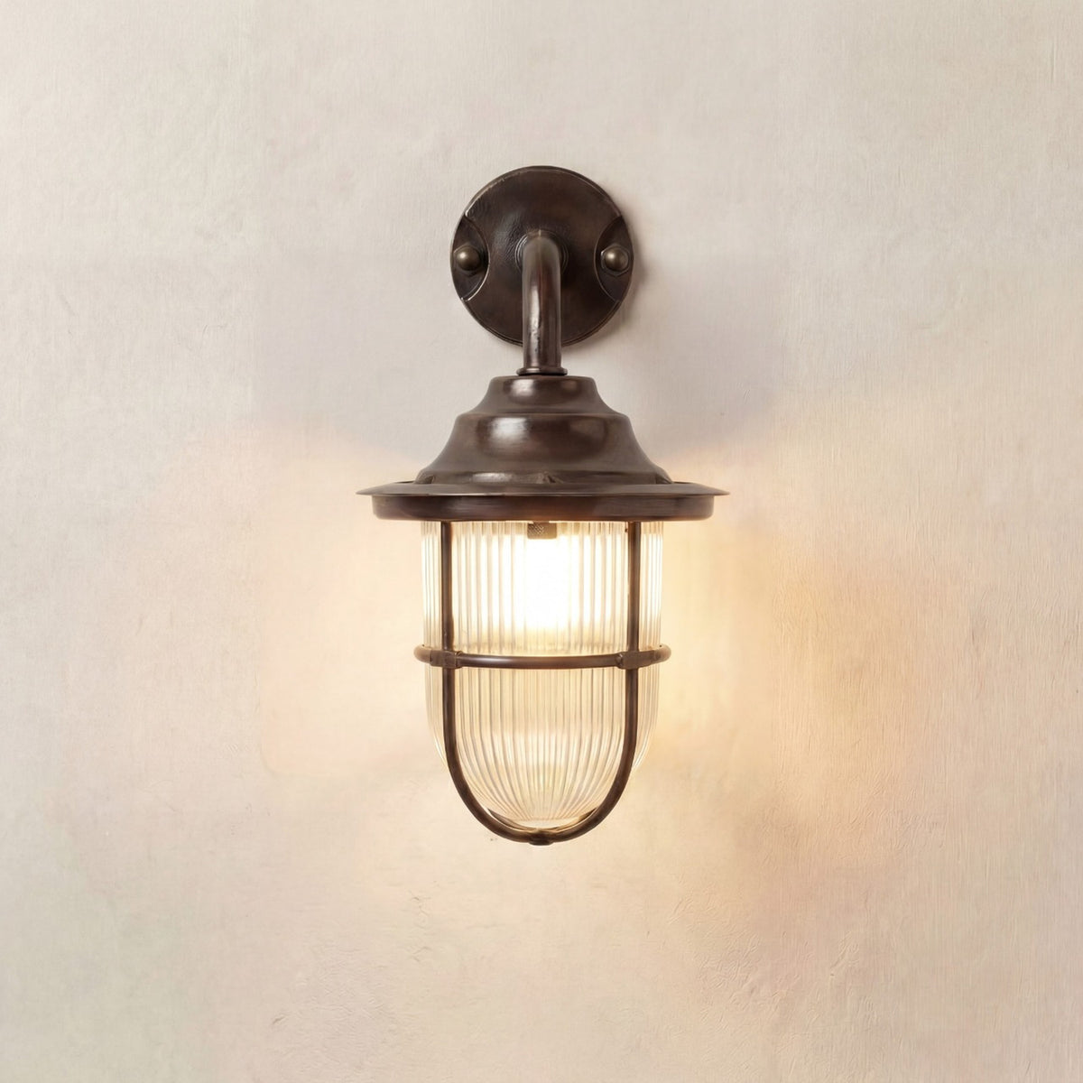 Quinn Guard outdoor wall lantern in antique brass with goose-neck arm, wide hood and ribbed glass shade in a cage guard, lit against a plain light background.