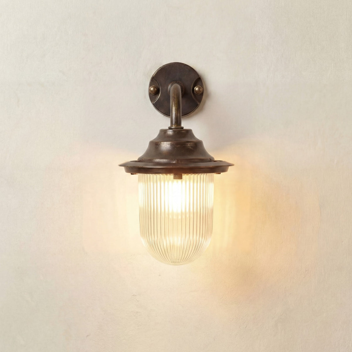 Quinn Outdoor Wall Lantern in antique brass, with a curved gooseneck arm, round wall plate, and ribbed glass shade lit against a plain light background.