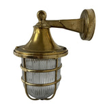 Grand Tiller outdoor wall lantern in raw brass, with flared hood, ribbed glass shade, brass cage guard and wall-mounted arm, shown on a plain white background.