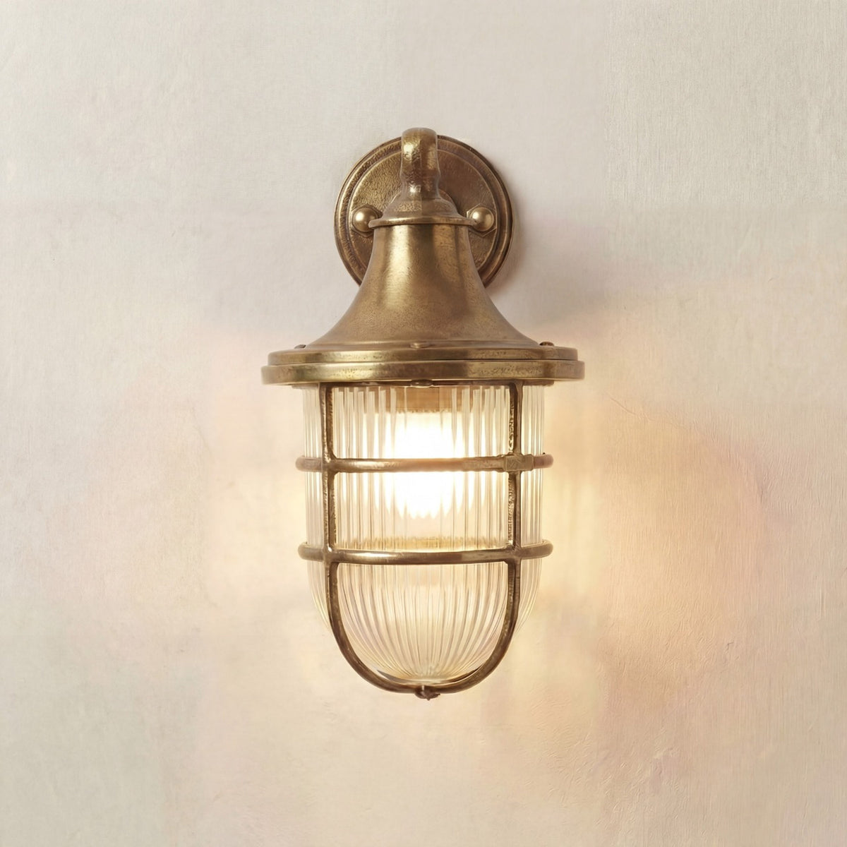 Grand Tiller outdoor wall lantern in raw brass, with flared hood, ribbed glass shade and cage guard, lit on a plain light background, IP64 rated.