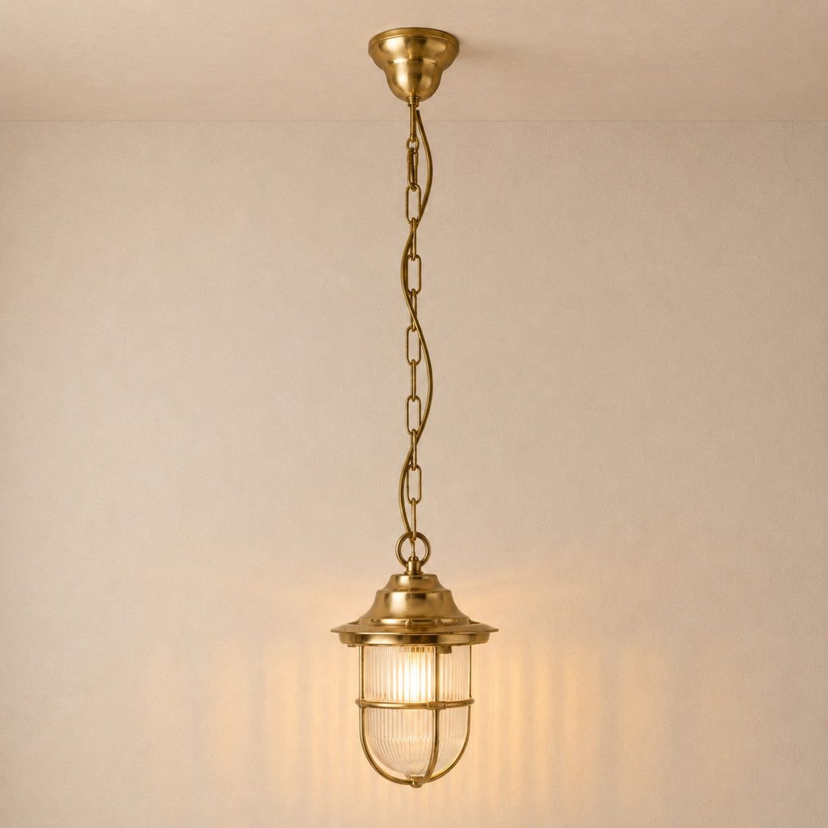 Hull Guard IP54 outdoor pendant light in raw brass, hung from a long chain with a lit ribbed glass shade inside a brass cage guard, against a plain light background.