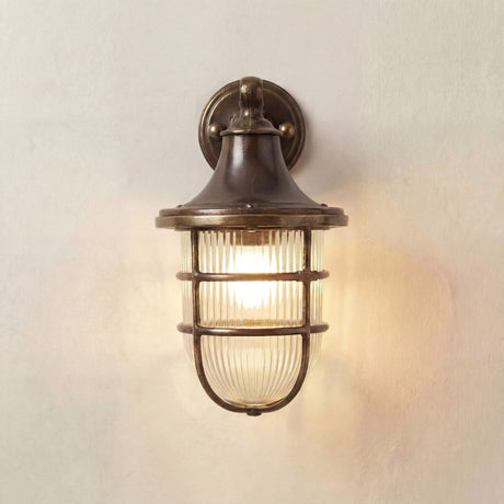 Grand Tiller IP64 outdoor wall lantern in antique brass, with bell-shaped hood, ribbed glass shade and brass cage, mounted on a short wall bracket against a plain light background.