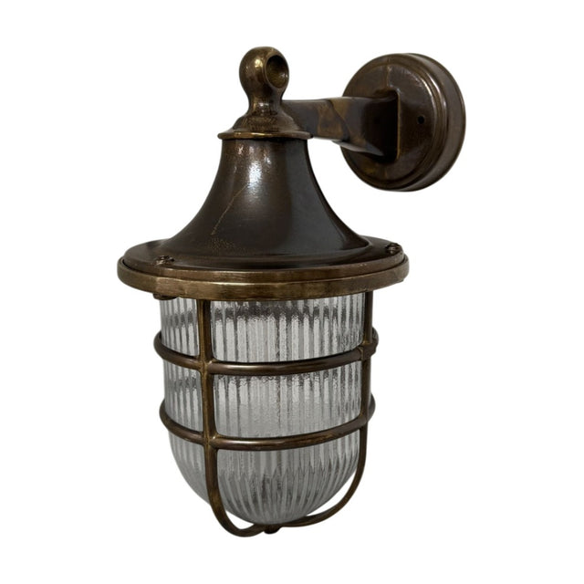 Grand Tiller outdoor wall lantern in antique brass finish, with bell-shaped hood, ribbed glass shade, and brass cage, mounted on a wall bracket against a plain white background.