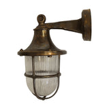 Tiller outdoor wall lantern in antique brass, solid brass bracket arm and round wall plate, bell-shaped hood over ribbed glass with cage guard, IP64, on a plain white background.