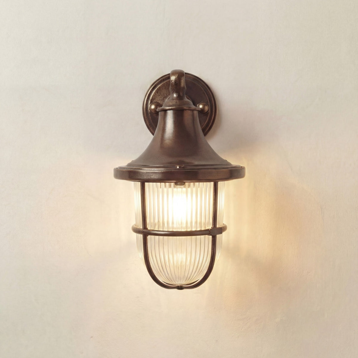 Tiller outdoor wall lantern in antique brass, with bell-shaped hood, ribbed glass, and cage guard on a round wall plate, lit against a plain light background.