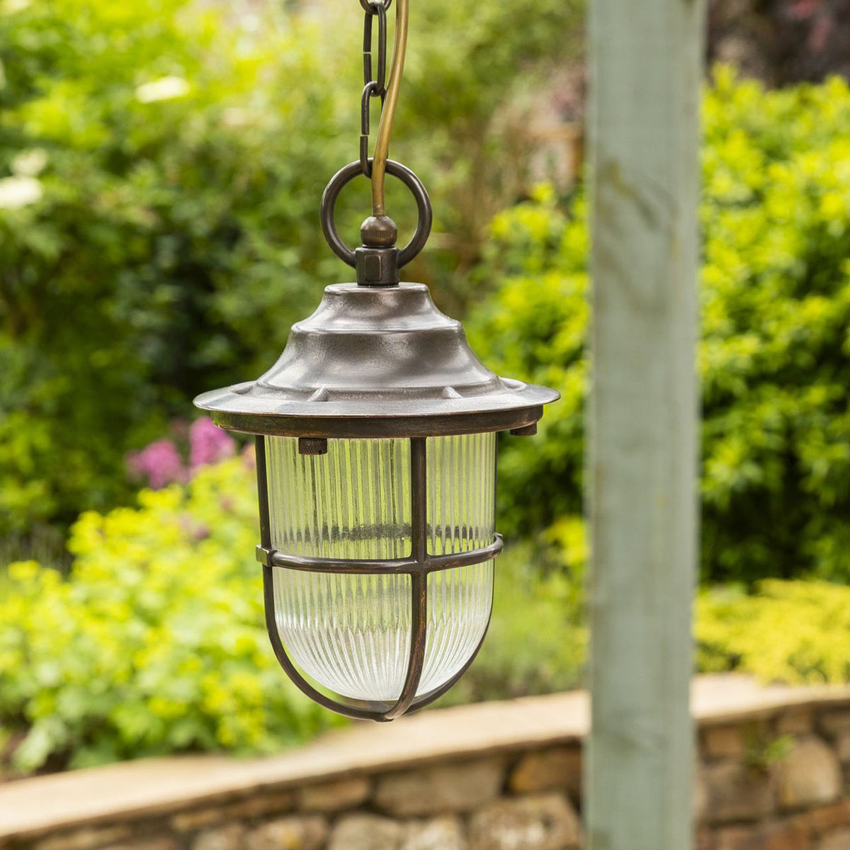 Hull Guard IP54 outdoor pendant light in antique brass, a caged lantern with ribbed glass shade and chain, shown hanging outdoors against a leafy garden backdrop.