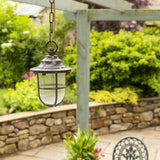 Hull Guard IP54 outdoor pendant lantern in antique brass with caged ribbed glass shade, hanging on a chain from a wooden pergola beam above a garden patio.