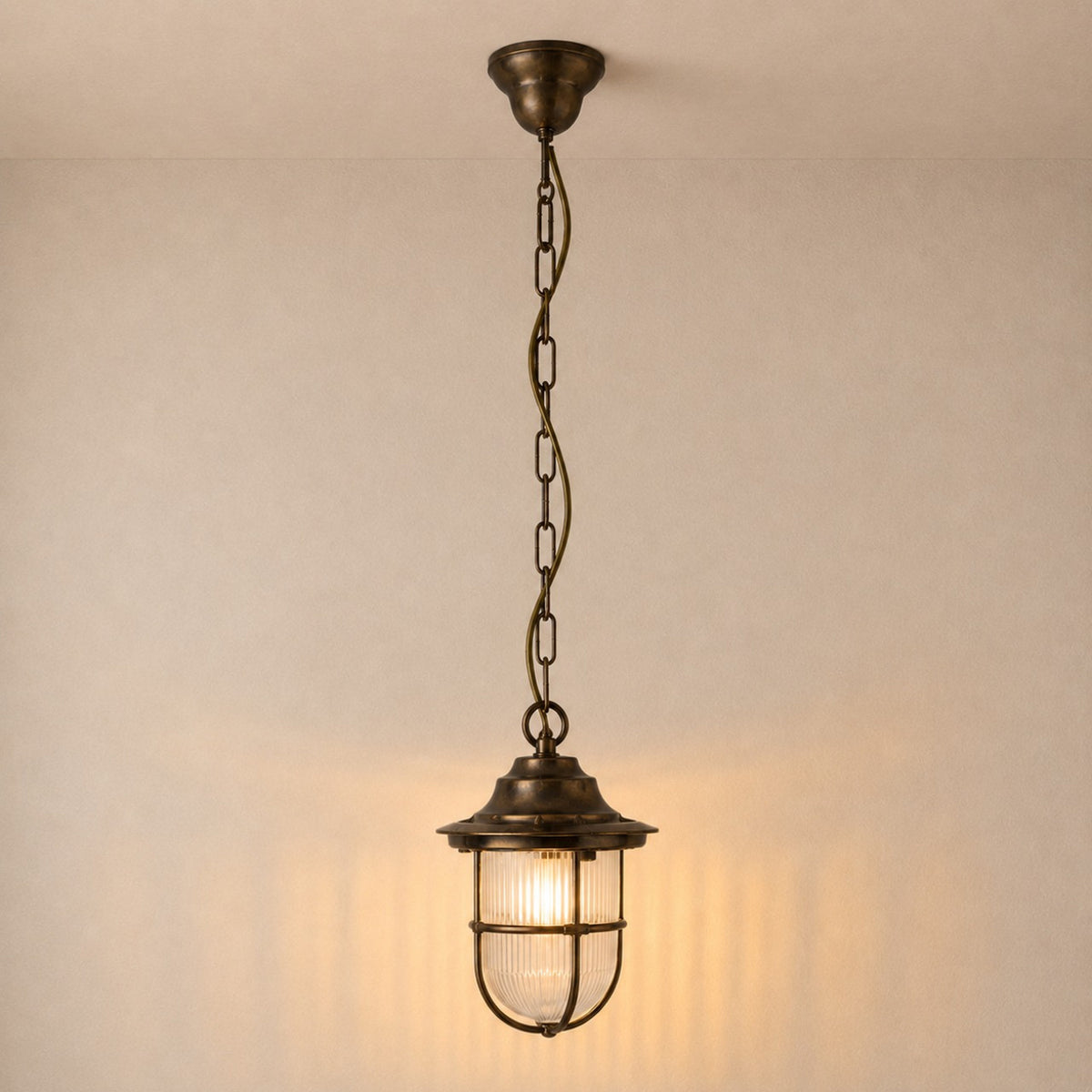 Hull Guard Outdoor Pendant Light in antique brass, a caged lantern with flared hood and ribbed glass shade on a long chain, shown illuminated against a plain wall.