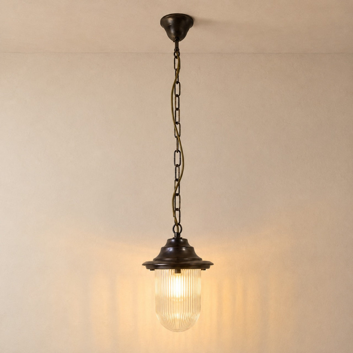Hull Outdoor Pendant Light IP54 in antique brass, with a flared hood over a clear ribbed glass shade, hung from a long chain and round ceiling rose against a plain light background.