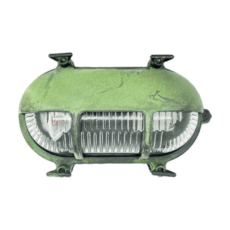 Palaemon eyelid bulkhead light in solid brass with verdigris green patina, oval body with hooded brow over ribbed glass divided by two bars, four corner mounting lugs, on white background.