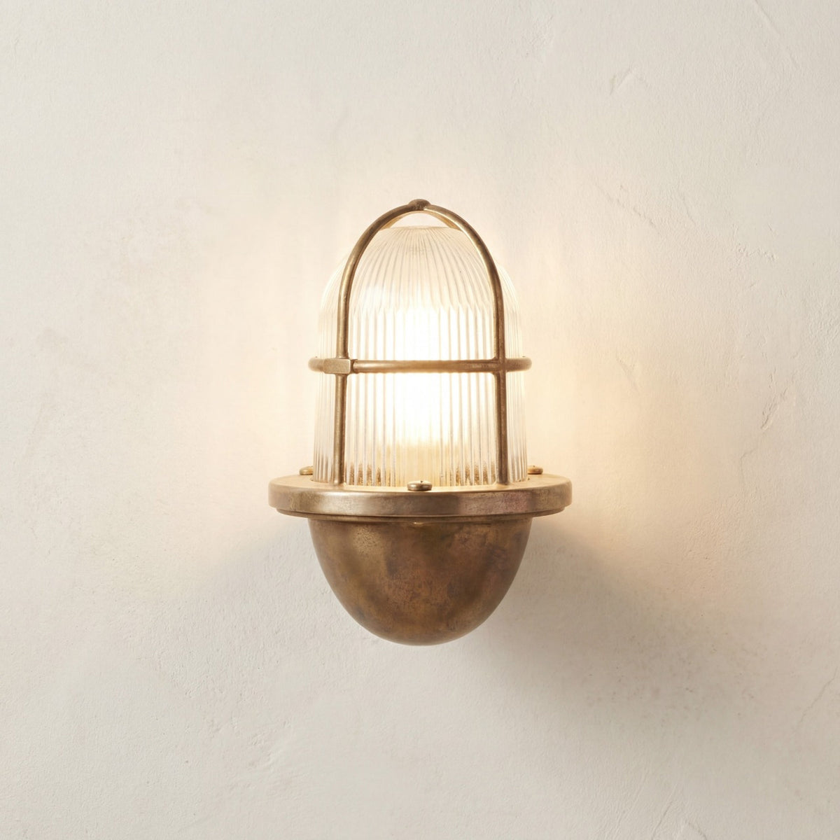 Armada Outdoor Wall Lantern in raw brass, lit uplight with ribbed glass dome and brass cage guard, rounded brass base, IP64 rated, shown on a plain light background.