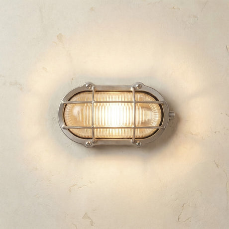 Merchant Oval Bulkhead Light in satin nickel, illuminated on a plain wall, showing its ribbed glass shade and protective cage in a nautical-style oval design.
