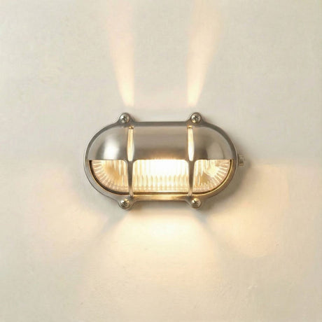 Palaemon North Oval Eyelid Bulkhead Light in satin nickel, wall-mounted with ribbed glass and crossbar guard, casting upward beams on a plain white wall.