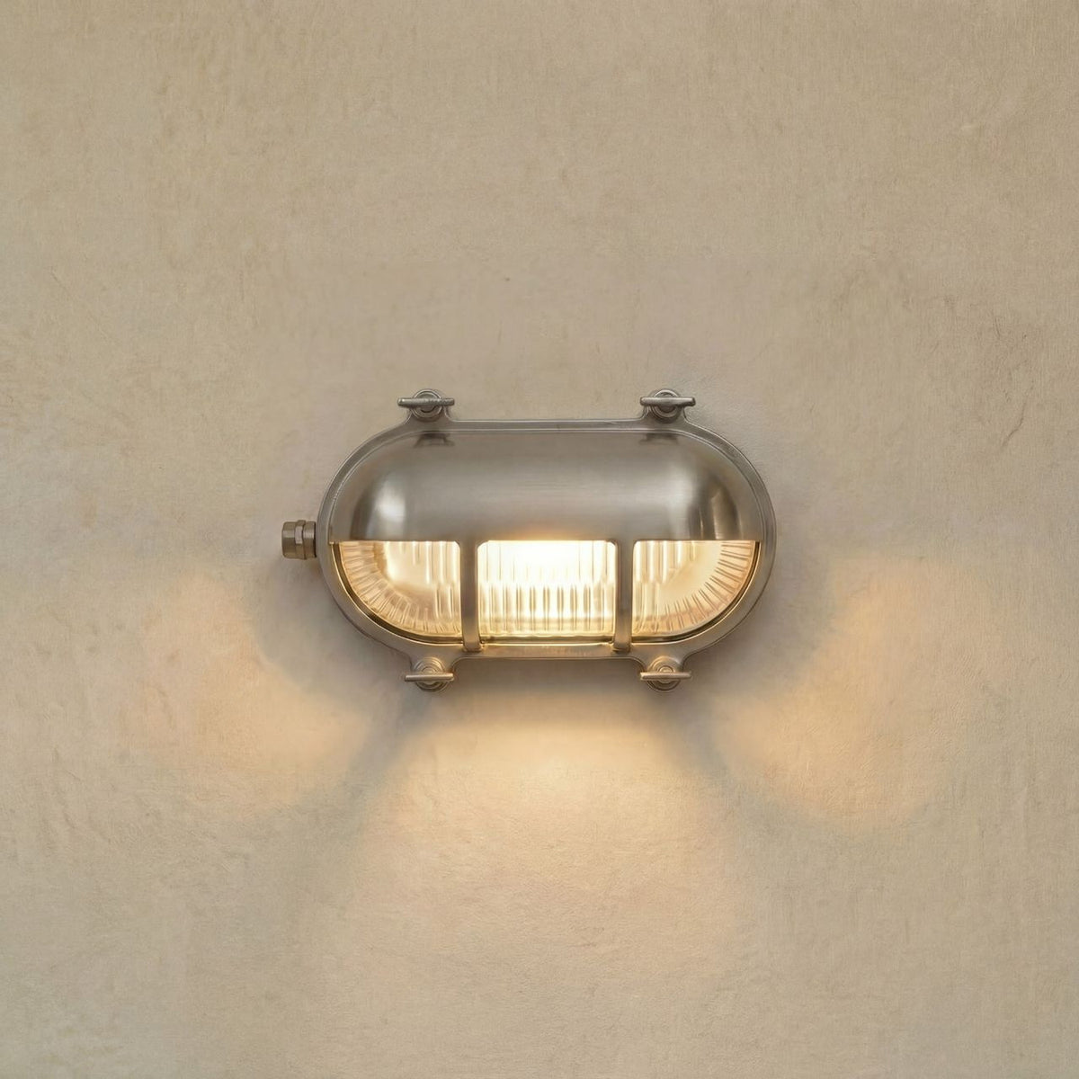 Palaemon Eyelid Bulkhead Light in satin nickel, an oval brass outdoor wall light with eyelid hood, ribbed glass and corner lugs, illuminated on a plain wall.