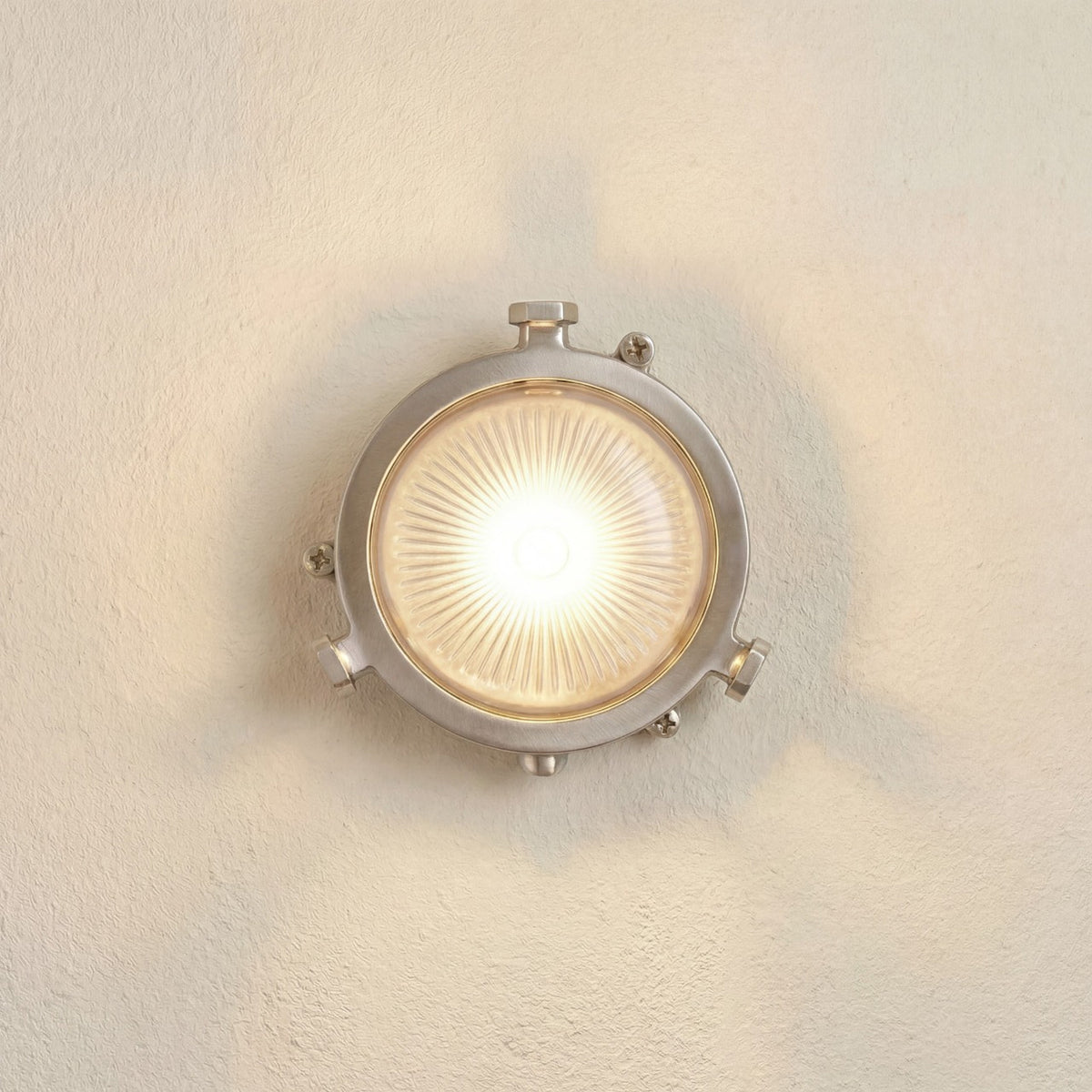 Mini Compass round bulkhead light in satin nickel, lit on a wall, showing a solid brass body, lugged rim, and frosted ribbed glass face.