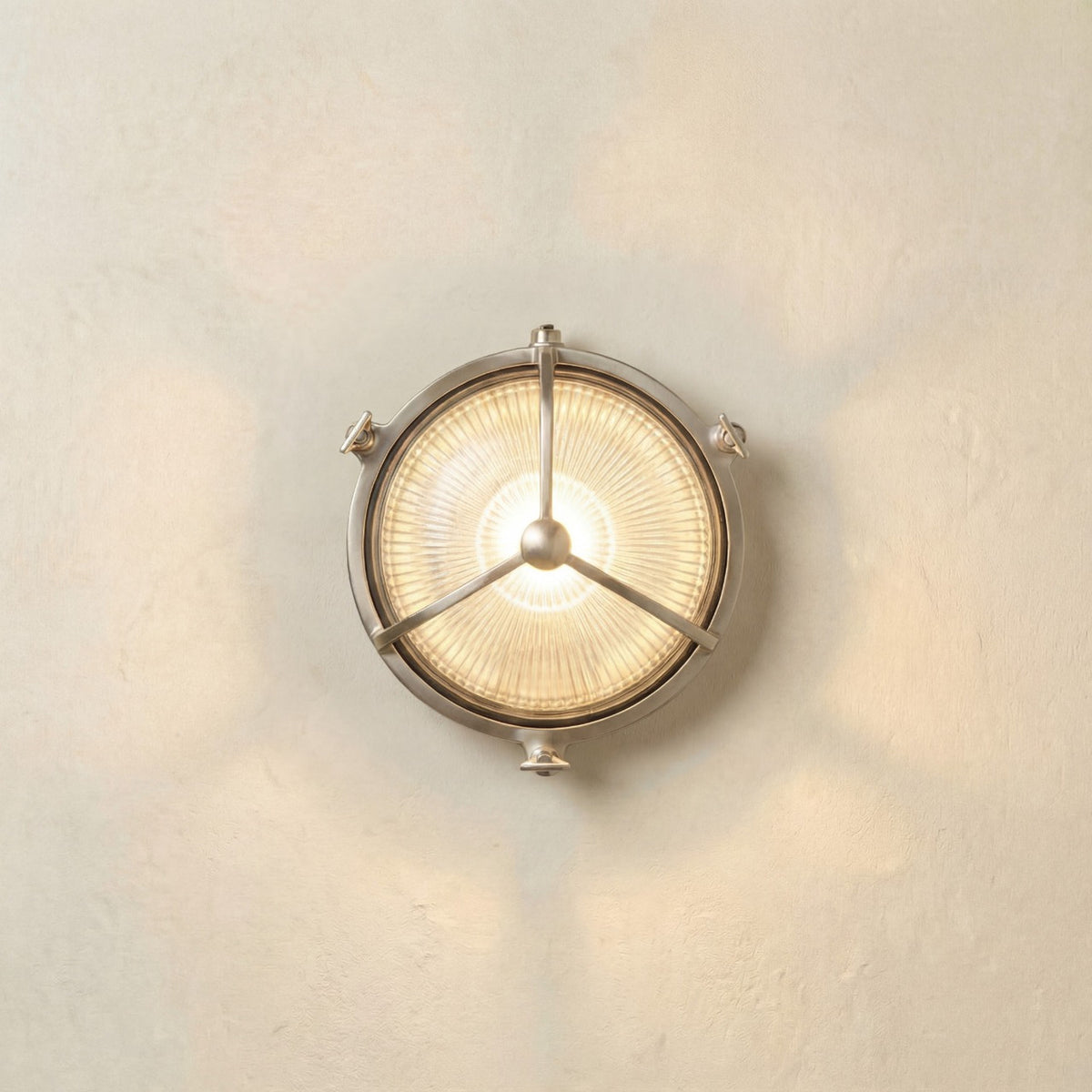 Trident Round Bulkhead Light in satin nickel, a brass outdoor wall light with ribbed glass and a three-arm guard meeting at a central domed boss, shown illuminated on a plain light wall.
