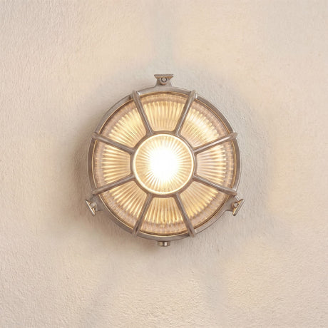 Grand Capstan round bulkhead light in satin nickel, with ribbed glass and radiating metal spokes, lit against a plain light wall, IP64 rated for outdoor use.