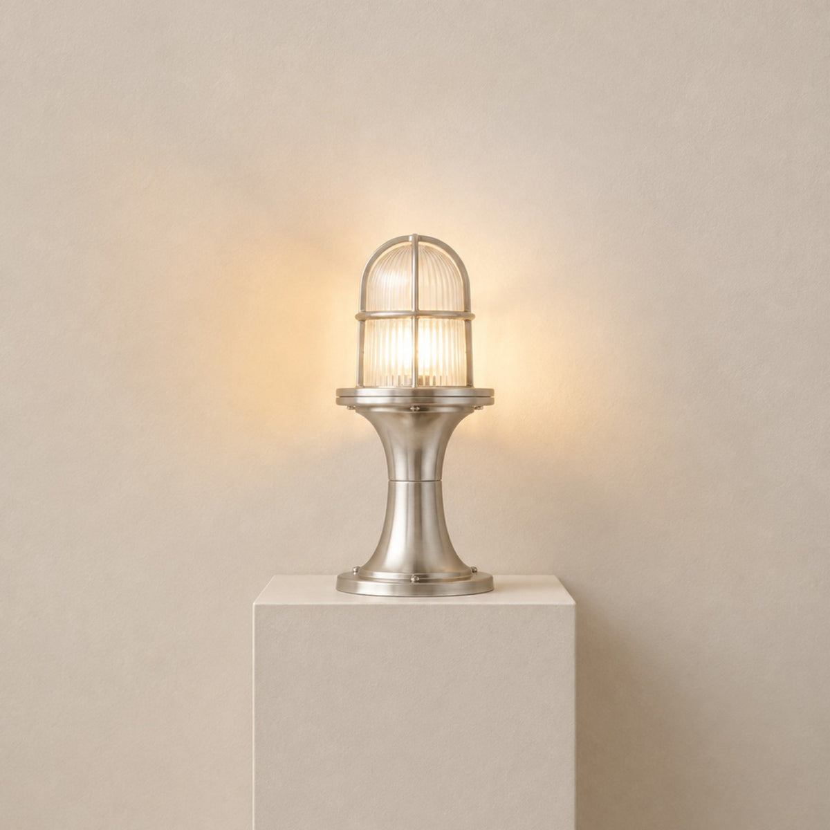 Wellington Outdoor Post Light IP54 in satin nickel, with a ribbed glass dome in a caged top on a waisted pedestal base, lit on a plinth against a plain light background.