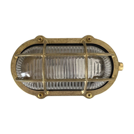 Merchant Oval Bulkhead Light in raw brass, IP64-rated, with a brass cage over ribbed glass and screw-fixed oval frame, shown on a plain white background.