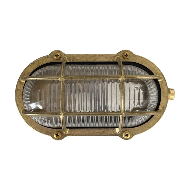 Merchant Oval Bulkhead Light in raw brass, IP64-rated, with a brass cage over ribbed glass and screw-fixed oval frame, shown on a plain white background.