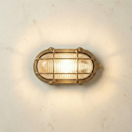 Merchant Oval Bulkhead Light in raw brass, an IP64-rated outdoor wall light with a brass cage over ribbed glass, lit and mounted on a plain textured wall.