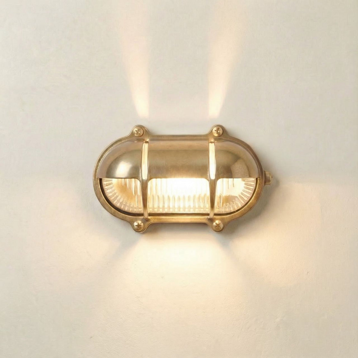 Palaemon North Oval Eyelid Bulkhead Light in raw brass, with ribbed glass lens and crossbar guard, illuminated on a plain white wall, casting light up and down.