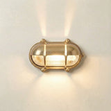 Palaemon North Oval Eyelid Bulkhead Light in raw brass, with ribbed glass lens and crossbar guard, illuminated on a plain white wall, casting light up and down.