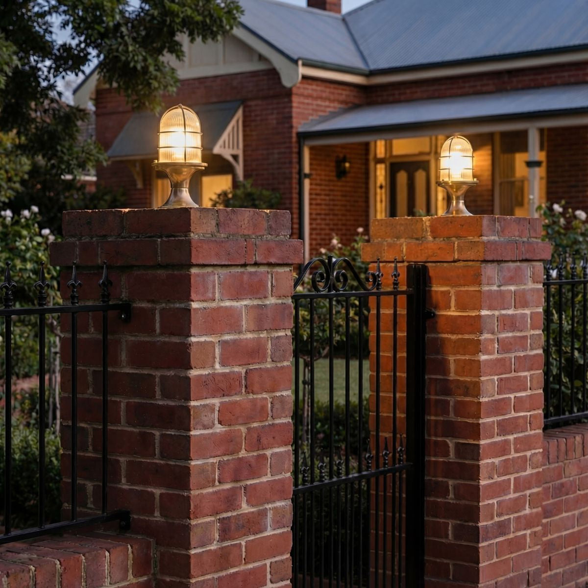 Wellington Grand IP54 outdoor post light in raw brass, caged ribbed glass dome on brick gate pillars either side of a red brick house entrance at dusk.