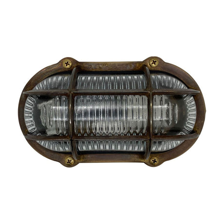 Merchant Oval Bulkhead Light in antique brass, with cross-head screws securing a protective cage over ribbed glass, IP64 rated, shown on a plain white background.