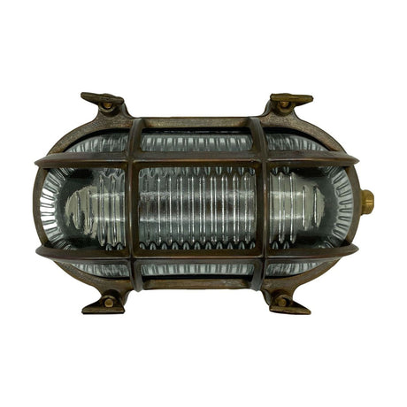 Horatio Junior oval bulkhead light in antique brass, with a crossed brass cage guarding ribbed glass and four wall-fixing lugs, shown on a plain white background.