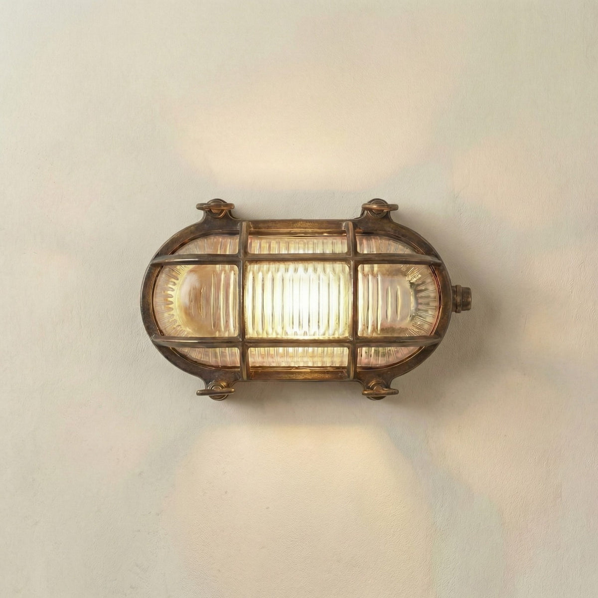 Horatio Junior oval bulkhead light in antique brass, with crossing brass cage bars over a ribbed glass diffuser, lit and mounted on a plain white wall.