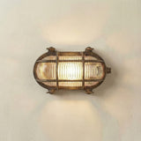 Horatio Junior oval bulkhead light in antique brass, with crossing brass cage bars over a ribbed glass diffuser, lit and mounted on a plain white wall.
