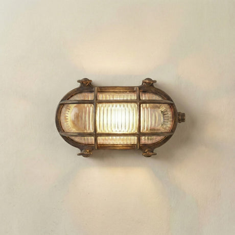 Horatio Junior oval bulkhead light in antique brass, with crossing brass cage bars over a ribbed glass diffuser, lit and mounted on a plain white wall.