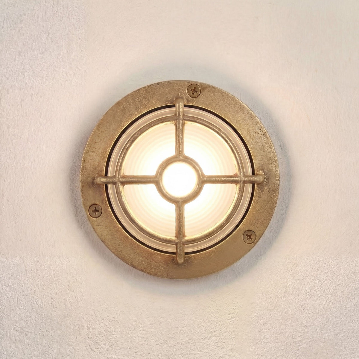 Walkway Guard outdoor wall light in raw brass, round flange with three screws, frosted glass lens, and a cross-bar brass guard, mounted on a plain white wall, illuminated.