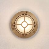 Walkway Guard outdoor wall light in raw brass, round flange with three screws, frosted glass lens, and a cross-bar brass guard, mounted on a plain white wall, illuminated.