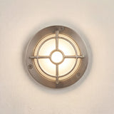 Walkway Guard IP65 outdoor wall light in satin nickel, round brass body with cross-and-ring guard over a glowing frosted glass diffuser, shown on a plain light wall.