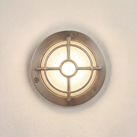 Walkway Guard IP65 outdoor wall light in satin nickel, round brass body with cross-and-ring guard over a glowing frosted glass diffuser, shown on a plain light wall.