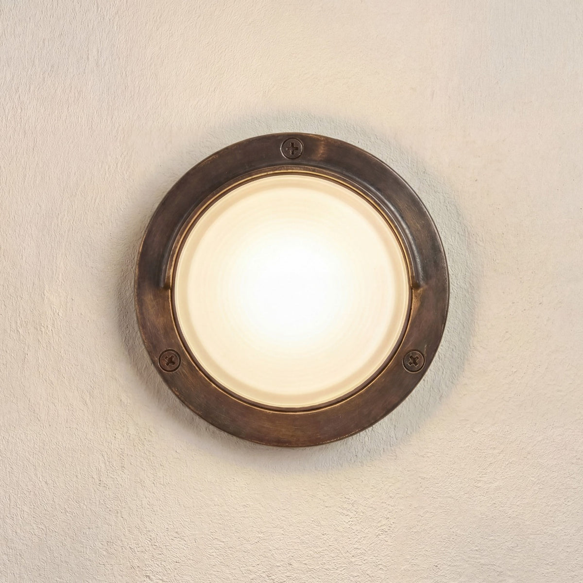 Walkway Eyelid Outdoor Wall Light in antique brass, round solid brass body with eyelid hood over a frosted glass lens, screw fixings visible, mounted on a plain beige wall.